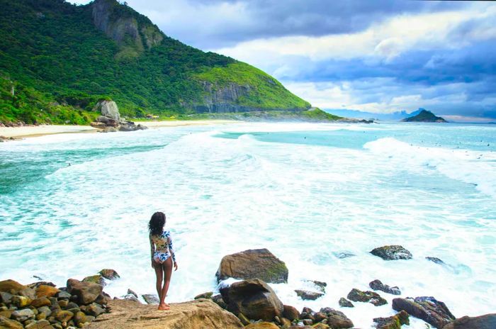 A young African-American woman in a one-piece swimsuit gazes out at a secluded beach beneath overcast skies.