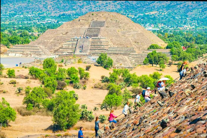 Visitors ascend the Pyramid of the Sun at the Teotihuacan pyramids, located near Mexico City, Mexico, on a bright sunny day, with the Pyramid of the Moon visible in the background.