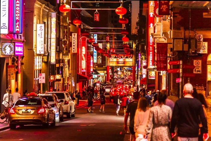 An evening scene in Chinatown, Melbourne, where people stroll through streets adorned with red lanterns hanging from buildings and arches above.