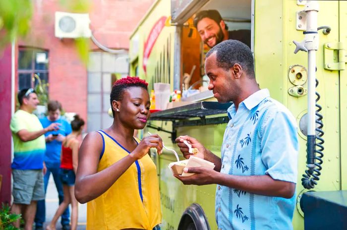 An African-American couple savoring their meal from a food truck on a lively city street, with people bustling around them. This horizontal waist-up shot offers ample copy space and was captured in Montreal, Canada.