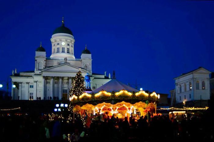 At night, a beautifully illuminated carousel spins at a Christmas market in Helsinki, set against the backdrop of the cathedral.