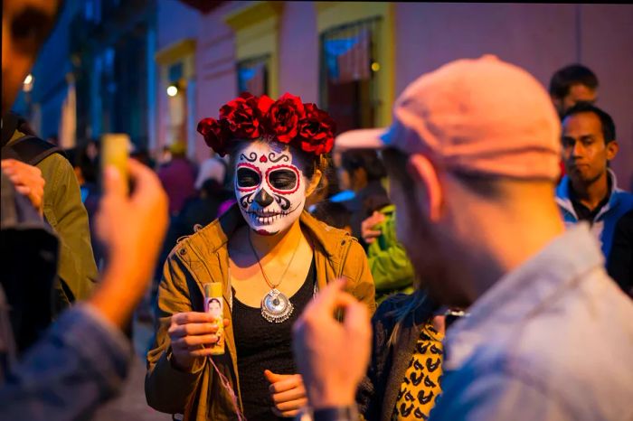 A young woman wearing a skeleton mask, clutching a drink while mingling with the crowd during Day of the Dead festivities in Oaxaca, Mexico.