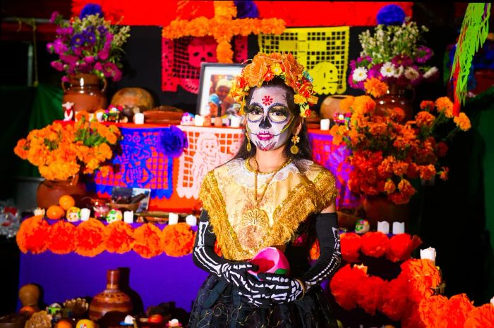 A girl in costume and face paint stands in front of an altar for the Day of the Dead festival.