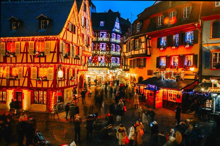 A historic town square, framed by medieval architecture, sparkles with Christmas lights as throngs of people move through the festive market.