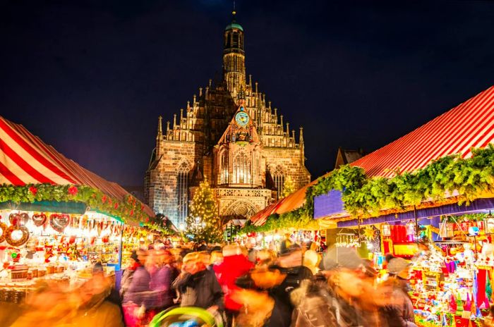 A large crowd moves through Nuremberg's famous Christmas market (Nuremberg Christkindlesmarkt) at night, surrounded by vibrant illuminated decorations and food stalls.