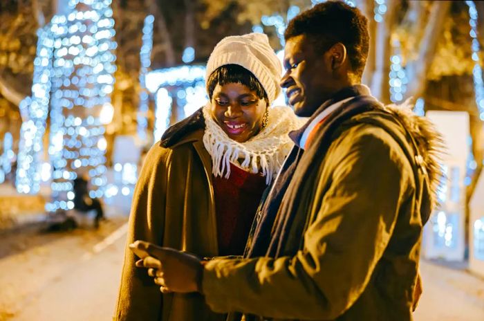 A young African American man shares his mobile phone with his mother in front of a beautifully lit Christmas tree at the Zagreb Christmas market at night.