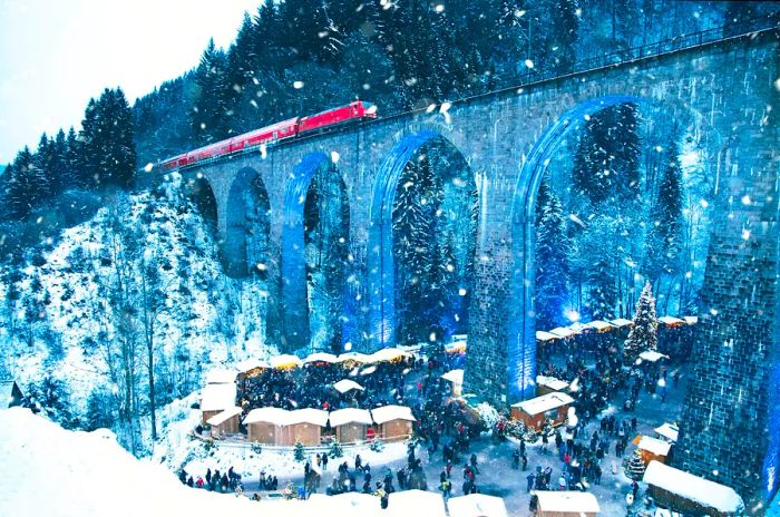 A picturesque Christmas market nestled in the Ravenna Gorge, Germany, set beneath a massive aqueduct as a train passes overhead amid falling snow.