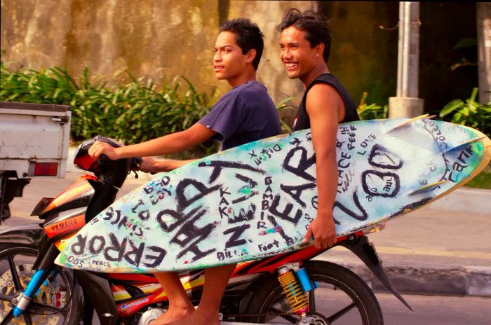 Two locals in Bali riding a motorbike with a surfboard.