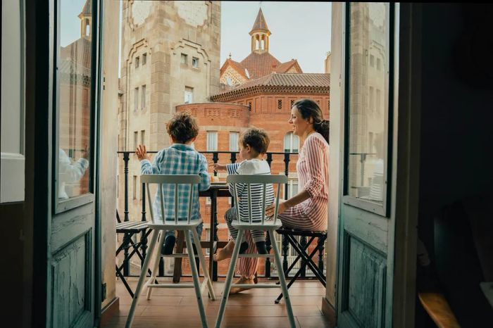 A mother and her two young children enjoy a moment on a balcony with a view of Barcelona's historic architecture.