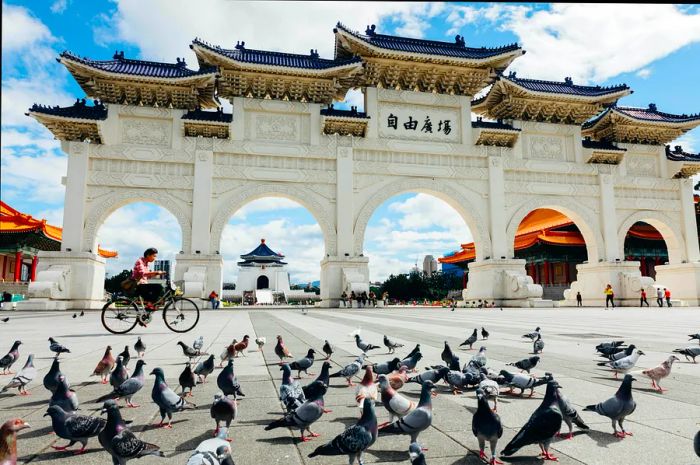 A woman rides her bicycle past the National Chiang Kai-shek Memorial Hall in Taipei on a sunny day, surrounded by pigeons roaming the square.