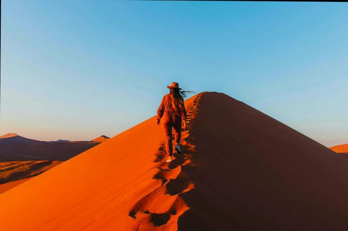 A young woman with long hair, wearing a hat and jumpsuit, ascends a towering dune to witness the breathtaking sunrise at the remote Namib-Naukluft National Park in Southern Africa, Namibia.