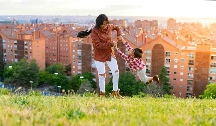 A mother joyfully spins her young son around in a spacious hilltop area with the cityscape in view