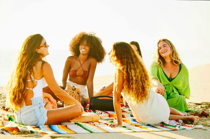 Two smiling female friends unwinding on a blanket at a tropical beach during sunset while vacationing in Mexico.