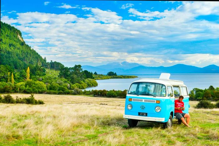 A man in a red shirt relaxes in the open side door of a turquoise VW camper van by Lake Taupo on New Zealand's North Island.