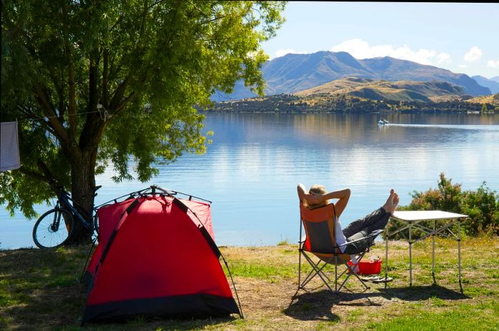 A woman enjoys a lakeside campsite, silhouetted against the sunset, reclining with her legs crossed and arms resting on her head, next to a tent and bicycle.