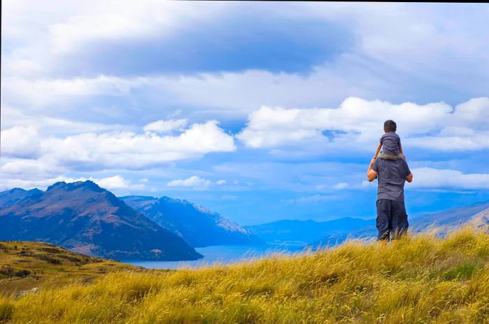 A child enjoys a scenic view of the sea and mountains in New Zealand while perched on his father's shoulders