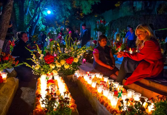 Two women sit beside candle-lit graves at night in Oaxaca, Mexico, while others admire the beautifully decorated tombstones in the background.