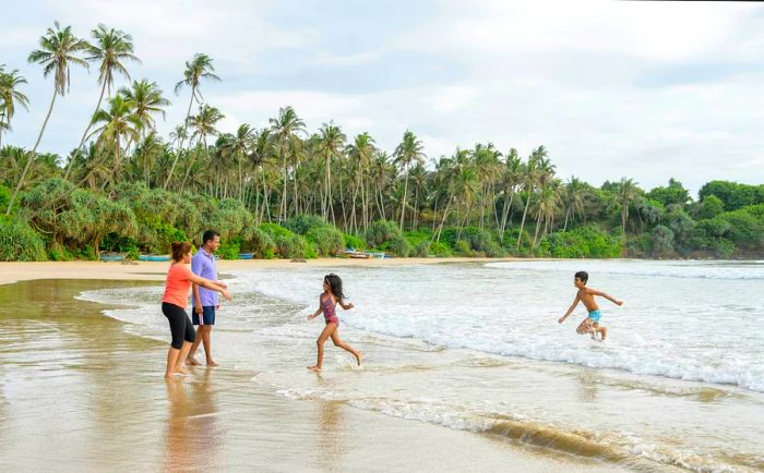 Two young children run from the sea towards their parents on a sandy beach lined with palm trees