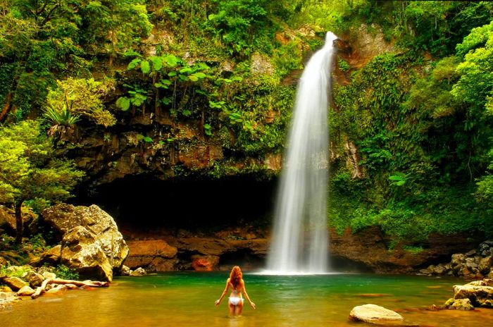 A woman stands in a tranquil pool before a cascading waterfall