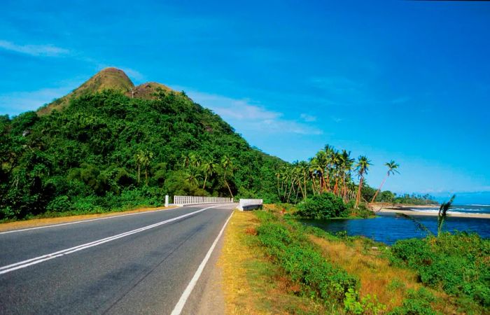 A deserted coastal road with a sandy palm-fringed beach on one side and a hill in the distance on Viti Levu, Fiji