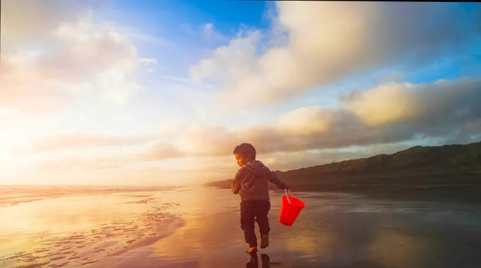 A young child runs along an empty beach near Auckland, clutching a red bucket as the sun sets.