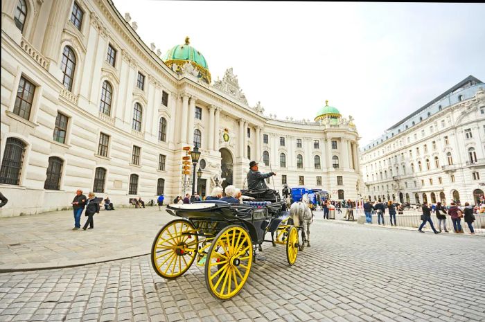 A horse-drawn carriage, known as a fiaker, glides past the Hofburg Palace at Michaelerplatz in Vienna, Austria