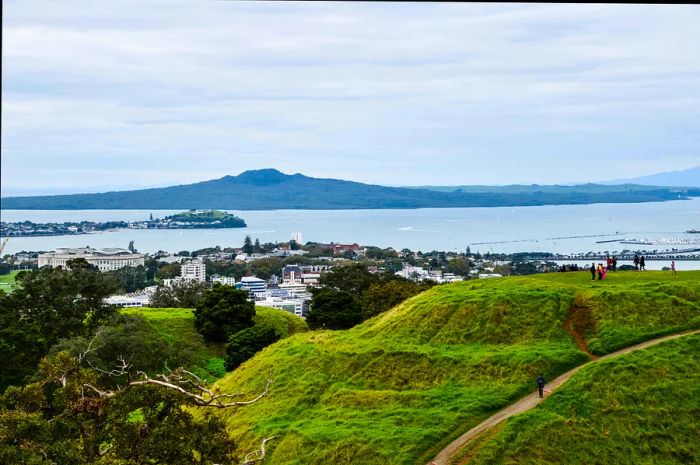 A grassy volcanic hill with hikers surrounding it at the edge of a bay