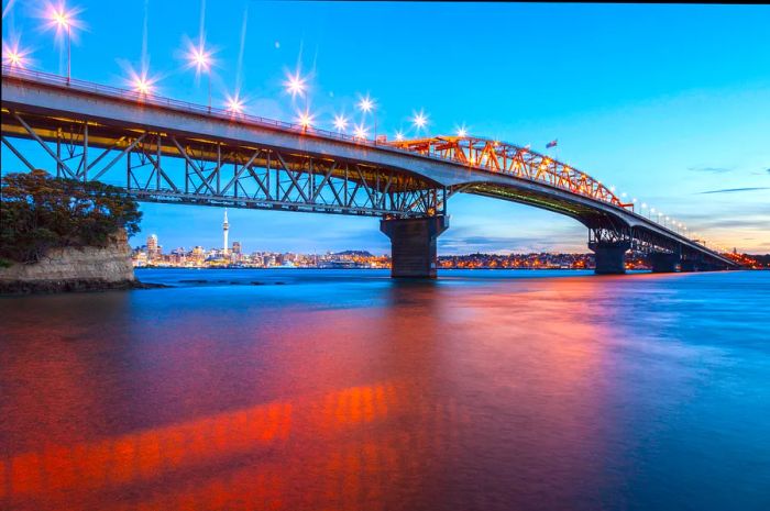 Auckland Harbour Bridge at dusk viewed from Northcote Point, with soft pink hues shimmering on the water's surface.