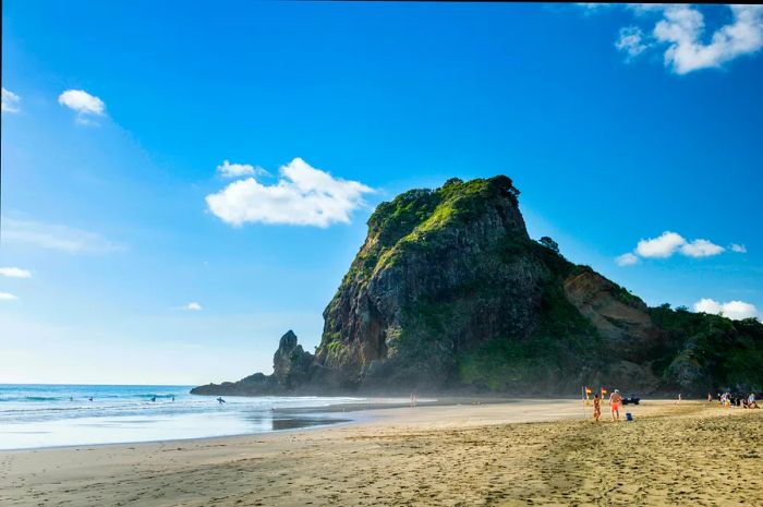 Swimmers and surfers make their way into the water, guided by flags that indicate the safe swimming area, with a towering rocky outcrop nearby.