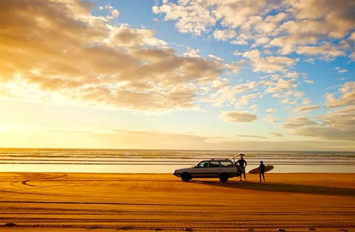 A vehicle parked on a sandy shore as two surfers prepare to hit the waves