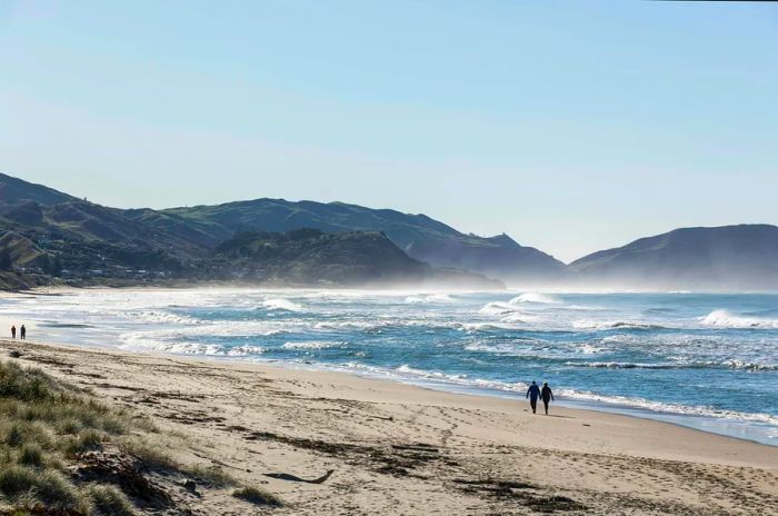 Silhouetted figures stroll along a beach with towering waves