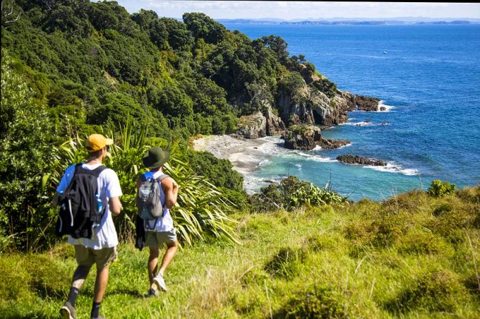 Two hikers with backpacks walk along a grassy path leading to a sandy cove flanked by tall rocks on either side.