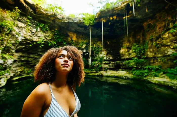 A woman stands by a cenote, a natural swimming hole, gazing up at the surrounding rock formations in Mexico.