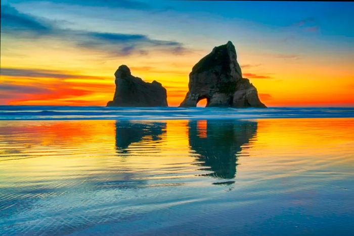 A crimson band stretches across the horizon as the sun sets behind the stone archways at Wharariki Beach in New Zealand