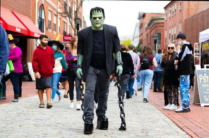 A large figure resembling Frankenstein's monster, painted white with a black outfit, strolls down the street.