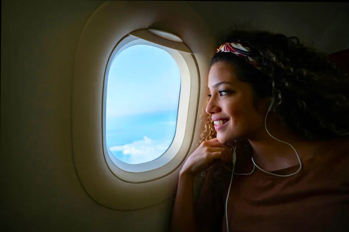 A joyful woman gazes out of an airplane window, enjoying music through her headphones.