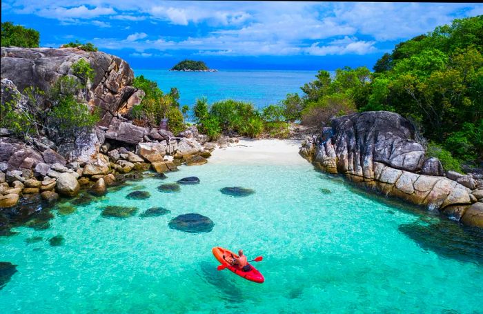 A man relaxes in a small red kayak, drifting through a turquoise bay surrounded by towering rocks.