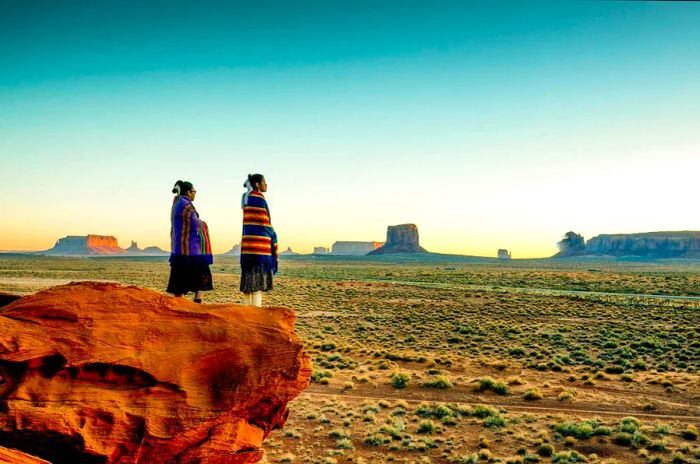 Two sisters in Navajo blankets stand on a rock at Monument Valley Tribal Park in the Navajo Nation