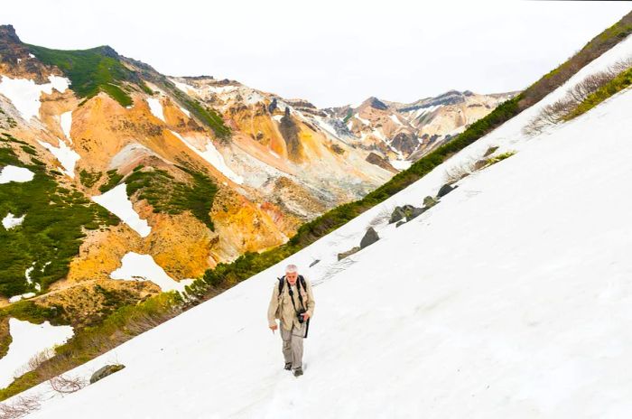 An older male hiker traverses a snow-laden slope with beige and brown volcanic hills in the background.