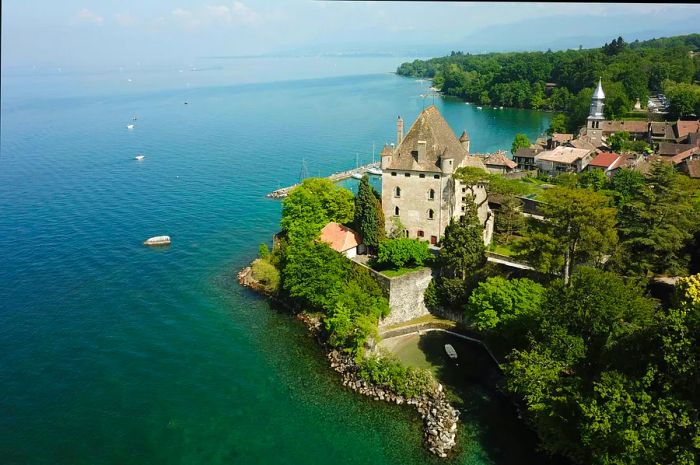 An aerial view showcases a stone castle perched on the shores of a stunning turquoise Lake Geneva