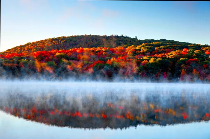 Mist envelops a lake in Connecticut, mirroring the vibrant reds, oranges, yellows, and greens of autumn leaves from a hillside above. A New England fall foliage road trip.