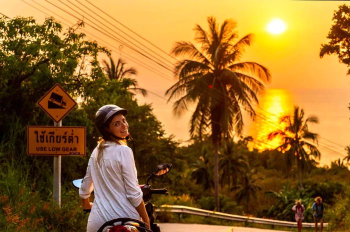 A woman wearing a helmet and sunglasses beams at the camera while riding a motorbike beneath palm trees in Thailand.