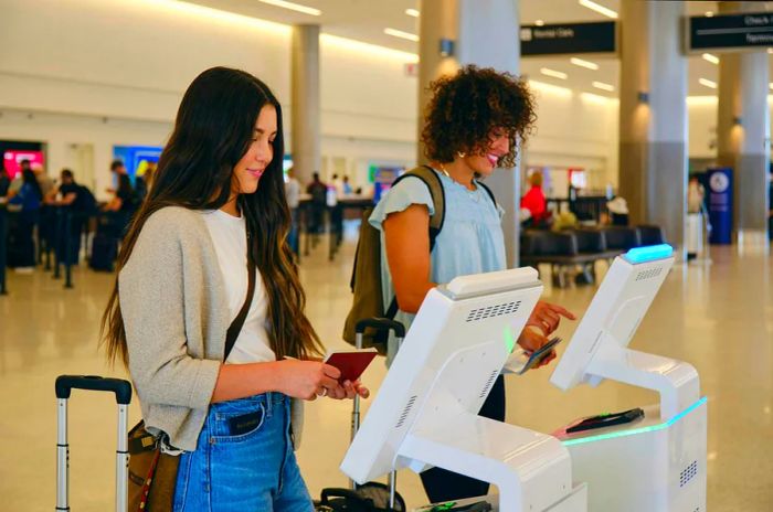 Two young women checking in at self-service kiosks in the airport.