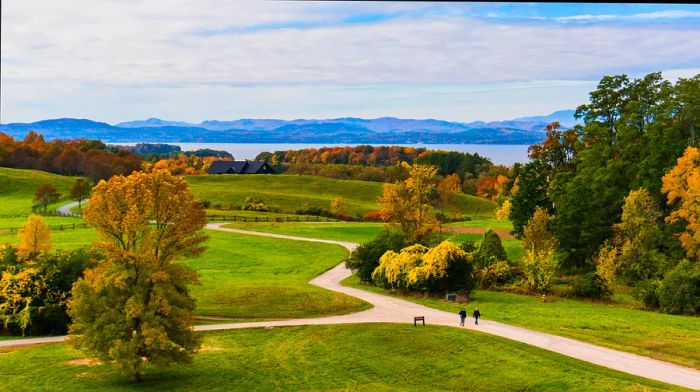 As the sun sets over Vermont, two individuals stroll along a trail at Shelburne Farms, with breathtaking views of Lake Champlain and the Adirondack Mountains in the backdrop.