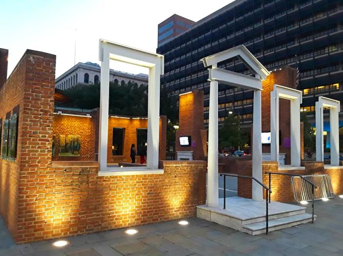 The exterior of a colonial-era home where George Washington once resided, now serving as an open-air museum, is illuminated at night, with a modern building in the background.