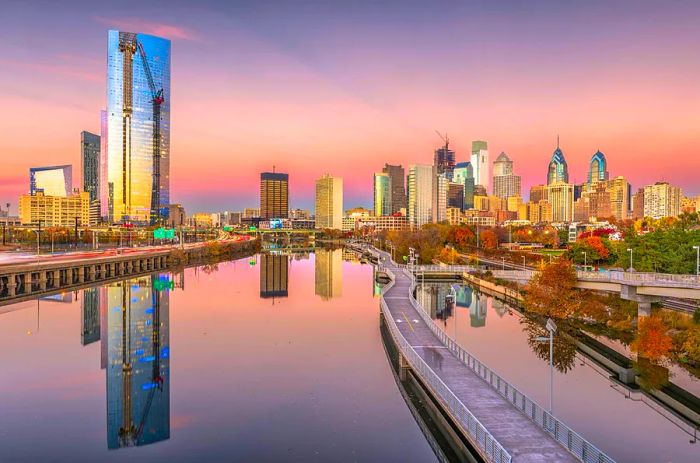 The skyline of Philadelphia, illuminated by a rose-pink and lavender sunset reflecting off the skyscrapers and the Schuylkill River, with the boardwalk trail in the foreground.