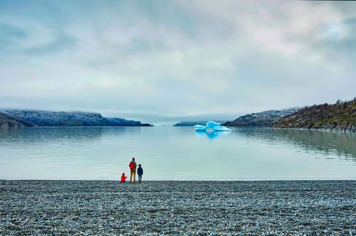 An adult stands with two young children at the edge of a lake, gazing at an iceberg.