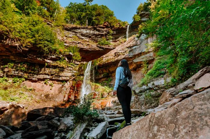 A woman stands on a rocky ledge, gazing up at Kaaterskill Falls.