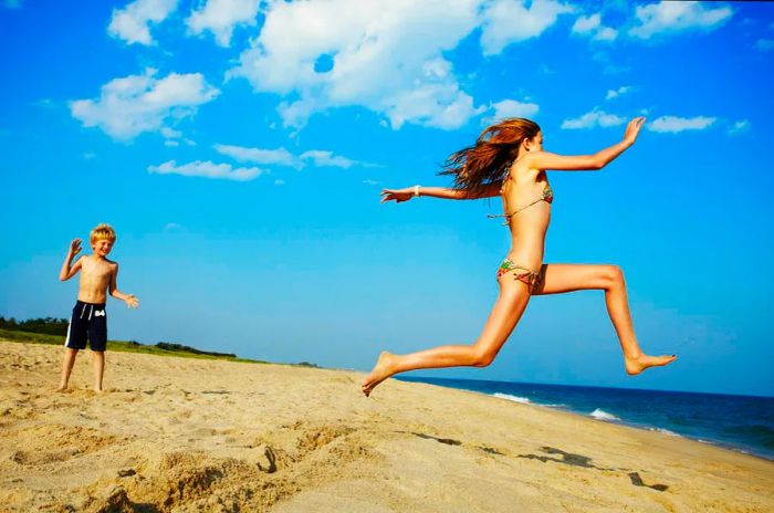 A girl joyfully runs along the pristine sands of the Hamptons in New York State, leaping toward the sea while a younger boy watches on.