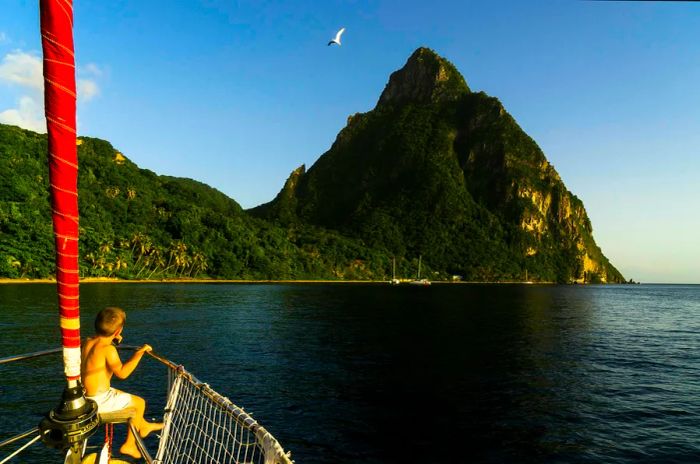 A young boy gazes out from the bow of a sailing boat, framed by the picturesque Piston Mountains in St. Lucia.